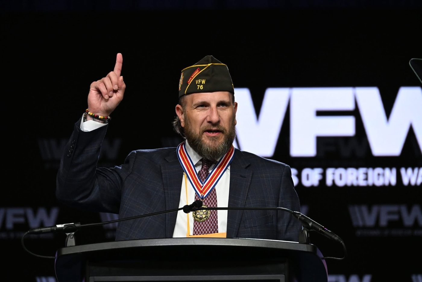 Tim Jensen speaking from a lectern in a VFW garrison cap, wearing the VFW Gold Medal of Merit on its ribbon, in front of a Veterans of Foreign Wars stage backdrop.
