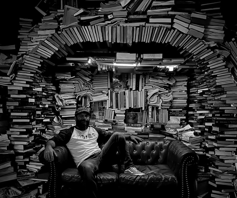 A man seated on a leather sofa inside an arched alcove built entirely from stacked books.