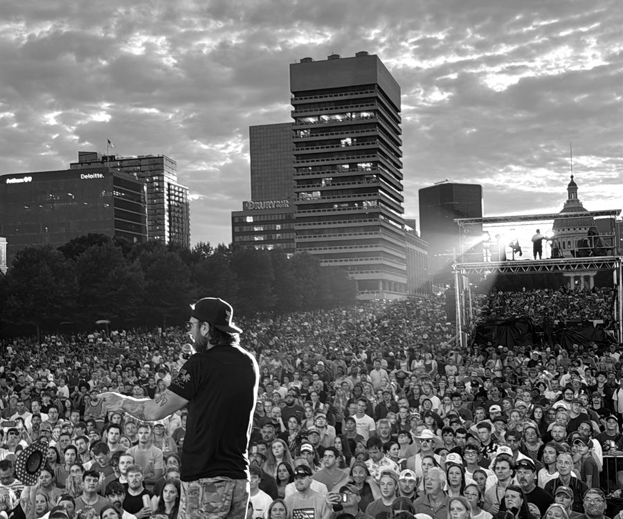 A man with a microphone addressing a large outdoor crowd at dusk, city skyline behind.
