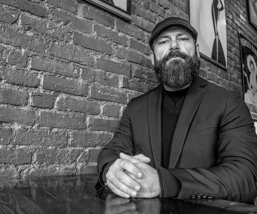 A bearded man in a flat cap and blazer seated at a table against an exposed brick wall.