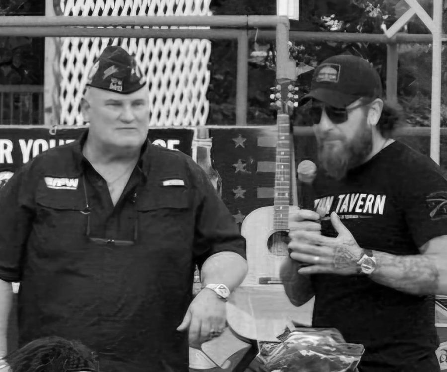 Two men on stage at a veterans community event, one in a VFW cap.